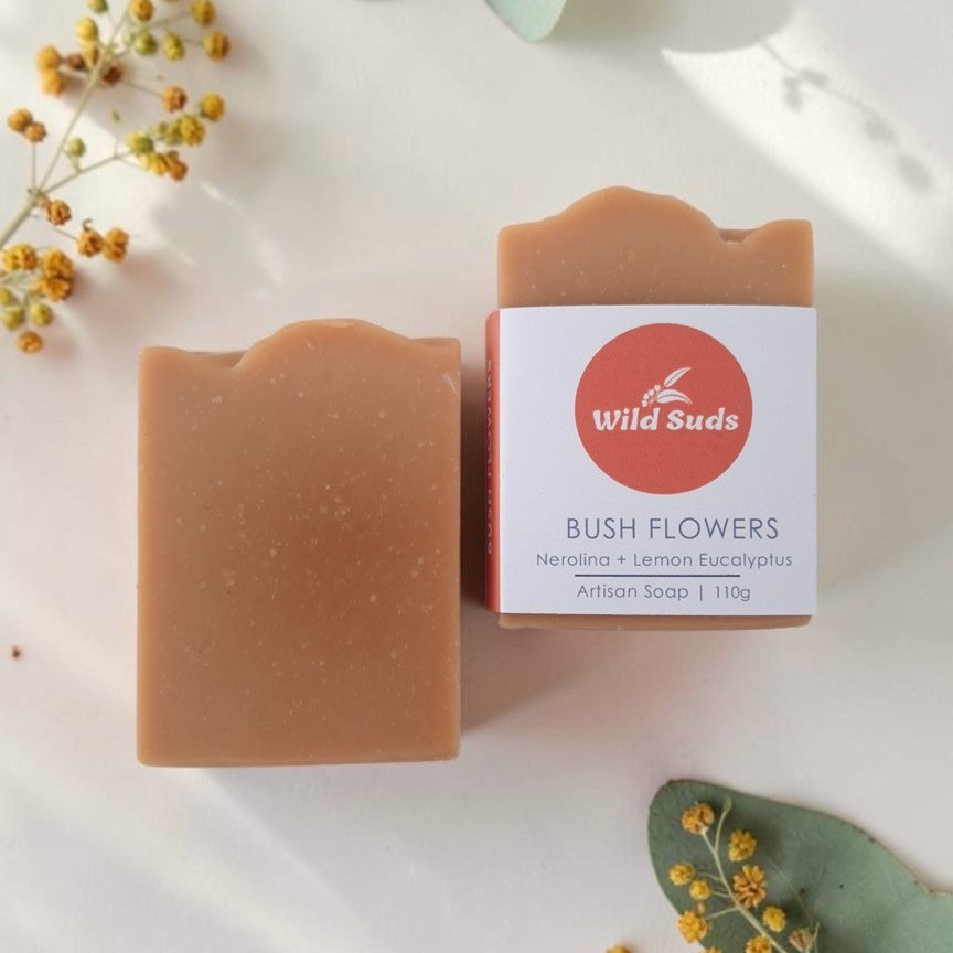 Two rectangular bars of Wild Suds 'Bush Flowers' handmade soap are displayed on a white background with decorative eucalyptus leaves and small yellow flowers. One bar is shown without packaging, revealing a smooth, speckled light brownish-orange surface.