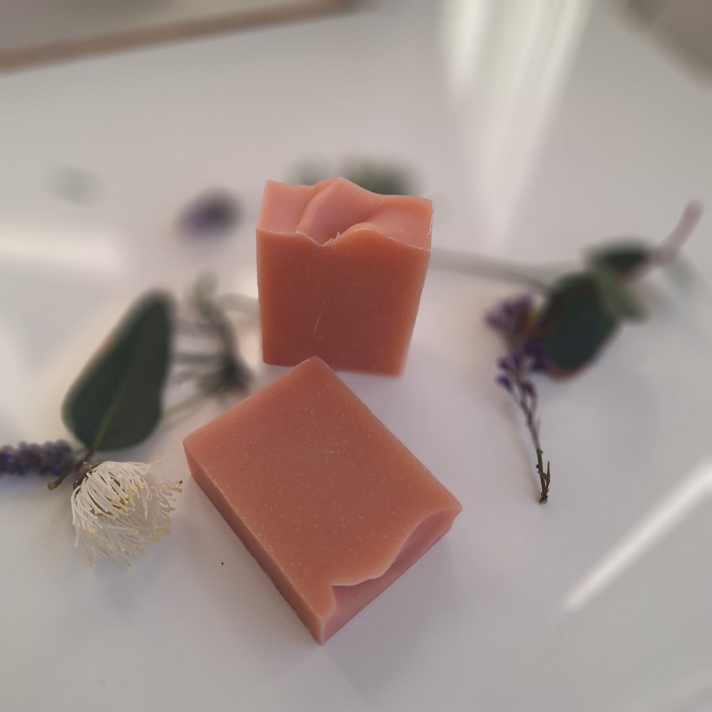 Two rectangular bars of dusty rose pink handmade soap with rustic, wavy cut edges, styled on a white surface with dried lavender, eucalyptus leaves, and a small white flower.