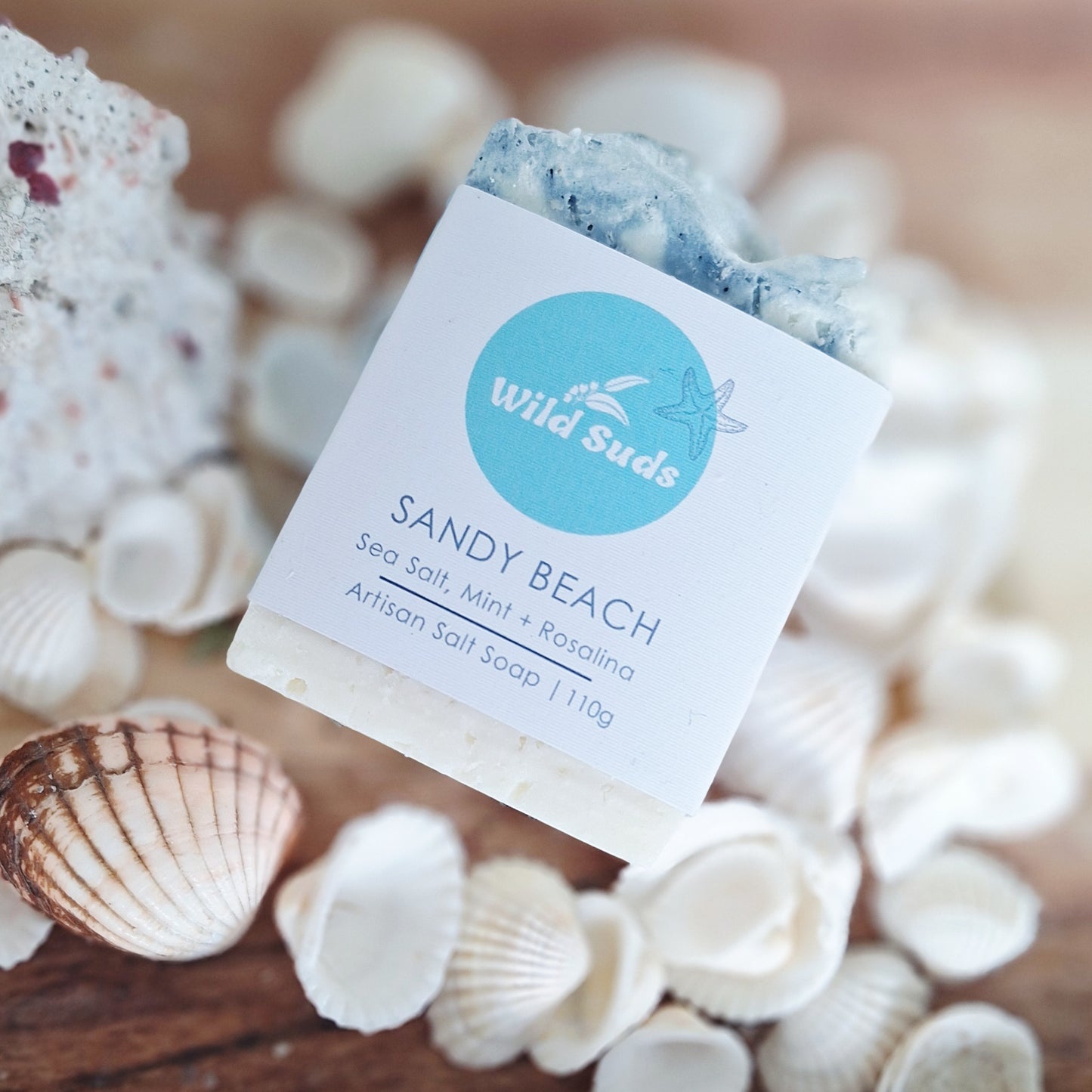 A bar of Wild Suds 'Sandy Beach' artisan salt soap, featuring a white paper label with a light blue circular logo, styled on a wooden surface surrounded by seashells. Scented with mint and rosalina
