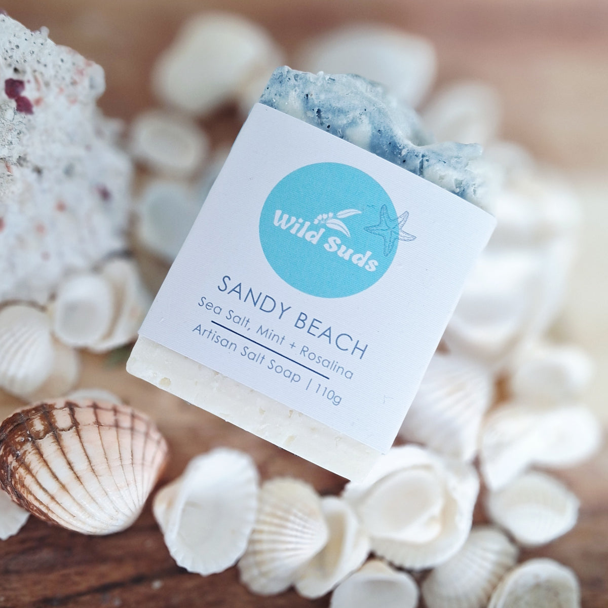 A bar of Wild Suds 'Sandy Beach' artisan salt soap, featuring a white paper label with a light blue circular logo, styled on a wooden surface surrounded by seashells. Scented with mint and rosalina