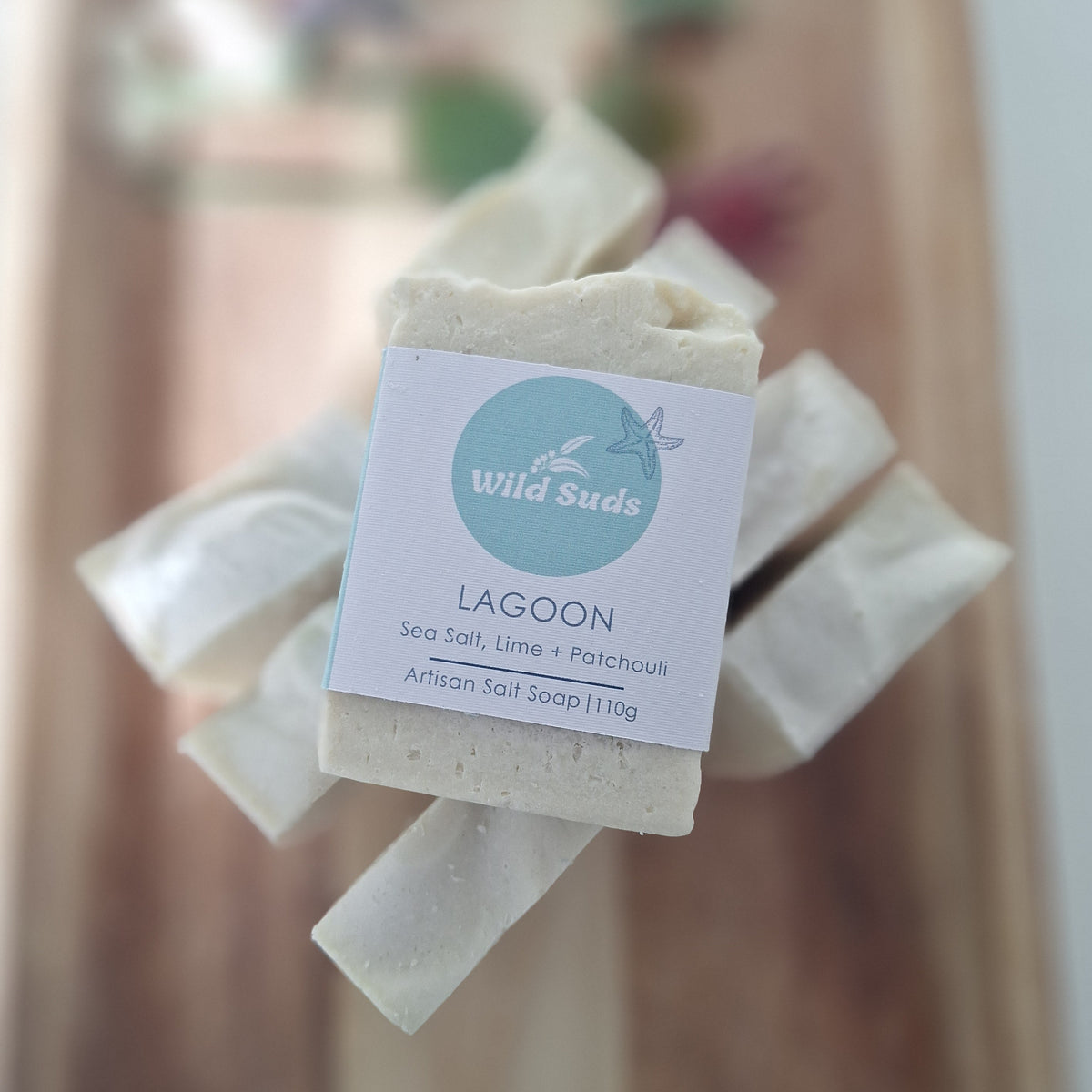 A handmade bar of soap labeled 'Wild Suds Lagoon' wrapped in kraft paper, held in front of a blurred wooden background.