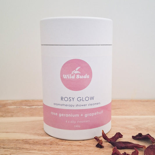 A white cylindrical container of Wild Suds Rosy Glow aromatherapy shower steamers, placed on a light wooden surface with dried rose petals in the foreground.