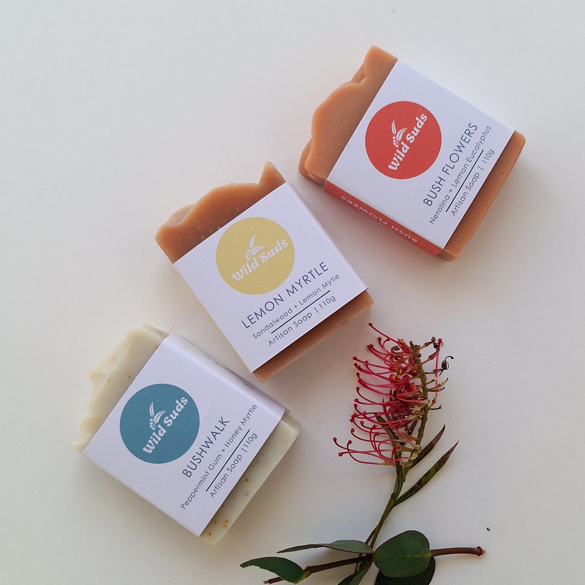 Three artisan soap bars from Wild Suds, labeled Bushwalk, Lemon Myrtle, and Bush Flowers, arranged on a clean white background with a small red flower stem as a decorative prop.