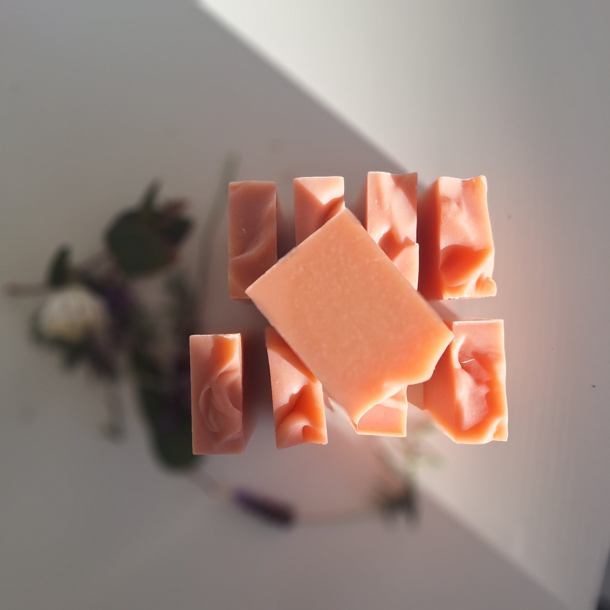 A set of peach-colored handmade soap bars arranged on a white surface with soft natural shadows and blurred dried flowers in the background.
