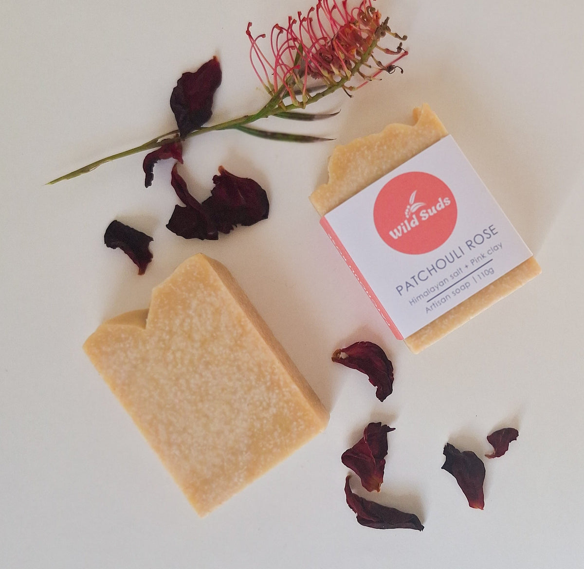 A flat-lay of a handmade soap bar next to its paper packaging wrapper, styled with dried rose petals and a wild flower on a clean white background. The wrapper is labele WILD SUDS and 'PATCHOULI ROSE'.